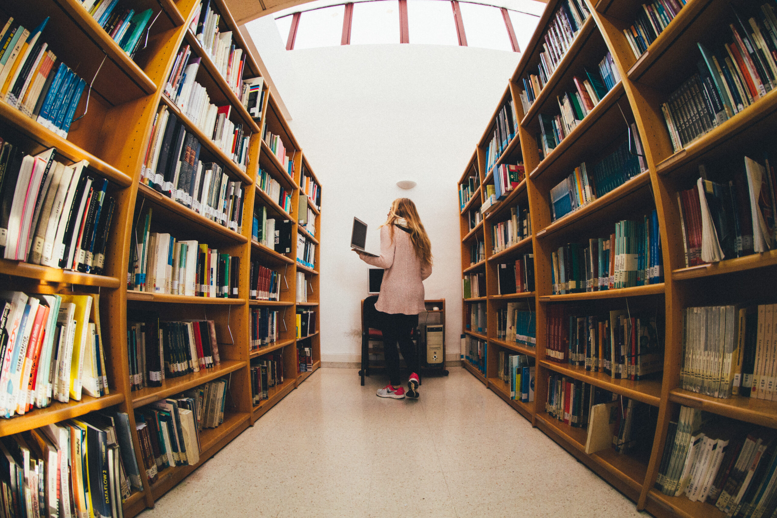 A women holding a laptop stands between library bookshelves looking at the books.